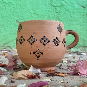 Peut inclure: Une tasse en terre cuite faite à la main, avec un corps arrondi et une anse incurvée. La tasse est ornée d'un motif répété de losanges noirs. La tasse est posée sur une surface en béton avec des feuilles et des pétales de fleurs tombées.