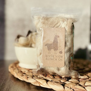 May include: A clear plastic bag filled with light brown alpaca fiber. The bag has a brown paper tag with a cartoon llama wearing a yarn ball and knitting needles. The tag reads "River View Alpaca Co."