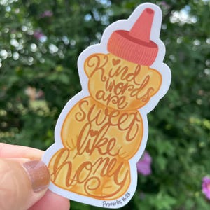 May include: A sticker in the shape of a honey bottle with the words "Kind words are sweet like honey" in orange script. The sticker has a light orange body and a red-orange cap. The text "Proverbs 16:23" is at the bottom.