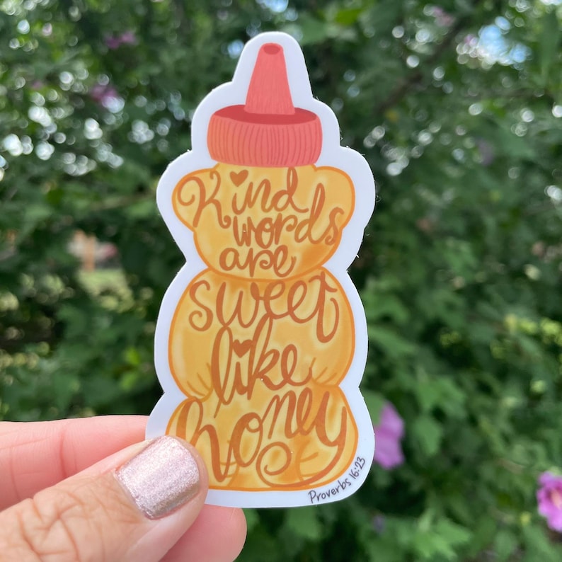 May include: A sticker in the shape of a honey bottle with the words "Kind words are sweet like honey" in orange script. The sticker has a white border and the text "Proverbs 16:23" in the bottom right corner. The honey bottle is orange and yellow.