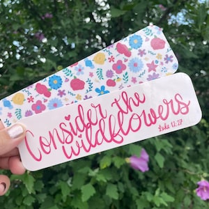 Könnte beinhalten: Zwei rechteckige Lesezeichen. Eines zeigt ein Blumenmuster mit rosa, blauen, gelben und lila Blumen auf weißem Hintergrund. Das andere Lesezeichen hat den Text "Consider the wildflowers" in rosa Schrift, mit "Luke 12:27" darunter.