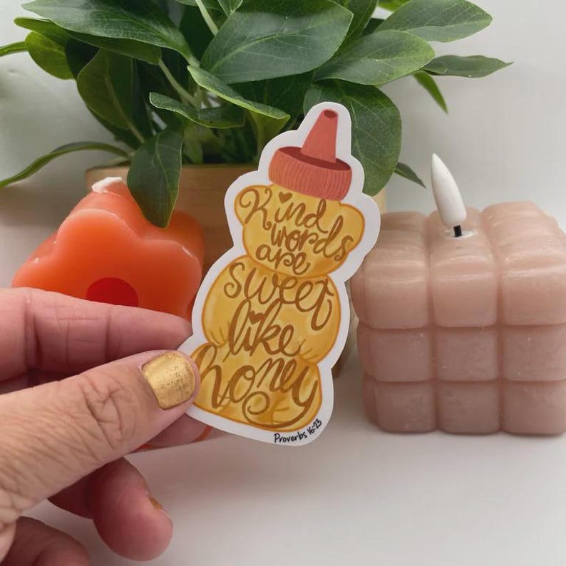 May include: A honey-shaped sticker with the text "Kind words are sweet like honey" in a cursive font. The sticker is held in front of a small orange candle, a pink cube candle, and a potted plant.