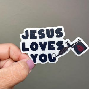 May include: A white sticker with the text "JESUS LOVES YOU" in bold, black letters. The sticker also features a tattoo gun and a vial of blood with a cross. The sticker is held by a hand.