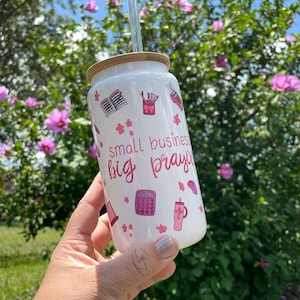 Puede incluir: Vaso de vidrio blanco con tapa de bambú y pajita transparente. El vaso está decorado con ilustraciones rosas de material de oficina y el texto "small business big prayers". El fondo es un arbusto verde con flores rosas.
