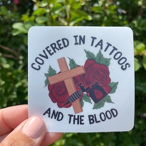 May include: Square sticker with the text "COVERED IN TATTOOS AND THE BLOOD". The design includes a wooden cross, red roses, and a tattoo machine. The sticker is held by a hand.