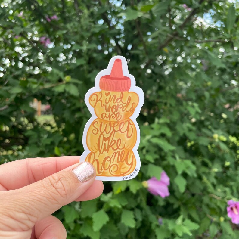May include: A sticker in the shape of a honey bottle with the words "Kind words are sweet like honey" in orange script. The sticker has a white border and a red-orange top. The background is a green bush.