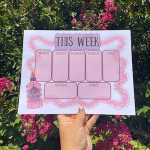 Könnte beinhalten: Ein Wochenplaner mit rosa und lila Design. Der Planer hat Abschnitte für jeden Wochentag, mit den Worten "This Week" oben. Der Planer enthält auch den Text "Jesus is the glitter glue holding my life together."