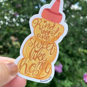 May include: A sticker in the shape of a honey bottle with the words "Kind words are sweet like honey" in brown script. The sticker has a light orange and red color scheme, with the text "Proverbs 16:23" at the bottom.