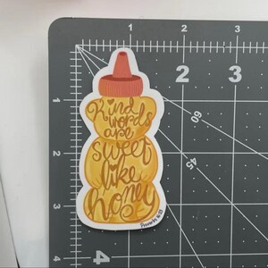 May include: A sticker in the shape of a honey bottle, with a brown cap and a yellow body. The sticker has the words "Kind words are sweet like honey" in a cursive font. The text "Proverbs 16:25" is at the bottom.