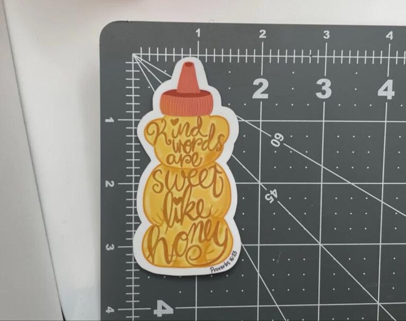 May include: A sticker in the shape of a honey bottle, with a brown cap and a yellow body. The sticker has the words "Kind words are sweet like honey" in a cursive font. The text "Proverbs 16:25" is at the bottom.