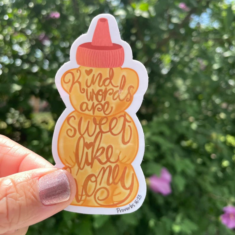May include: A sticker in the shape of a honey bottle with the words "Kind words are sweet like honey" in a handwritten style. The sticker has a light orange and brown color scheme, with a red cap. The text "Proverbs 16:23" is at the bottom.