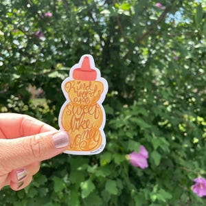 May include: A sticker in the shape of a honey bottle with the words "Kind words are sweet like honey" in orange script. The sticker has a white border and is held against a backdrop of green foliage and pink flowers.