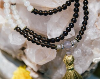 May include: A brown and white beaded mala necklace with a gold tassel. The necklace is resting on a bed of white quartz crystals.