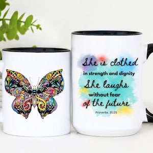 May include: Two white ceramic mugs with black rims. The mug on the left features a colorful butterfly design. The mug on the right has a watercolor background with the text "She is clothed in strength and dignity. She laughs without fear of the future. Proverbs 31:25."