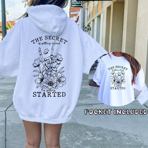 May include: White hoodie with a black graphic print on the back that says "The Secret of getting ahead is getting started" with a skeleton hand holding flowers. The front of the hoodie has a pocket.
