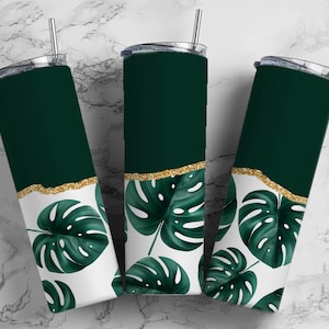 Könnte beinhalten: Drei Edelstahlbecher mit einem tropischen Blattdesign in Grün und Gold. Die Becher haben einen weißen Hintergrund mit grünen Monstera-Blättern und einem goldenen Glitzer-Akzent.