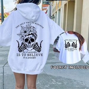 May include: White hoodie with a black skull and floral design on the back. The text reads "The only way to achieve the impossible is to believe it is possible".