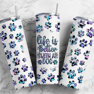以下が含まれることがあります： 白い背景に青、ピンク、黒の肉球模様が描かれたステンレス製のタンブラーが3つ。中央のタンブラーには、白いアウトラインで囲まれた黒い文字で「Life is better with a dog」と書かれている。