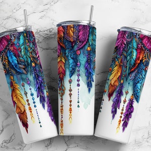 May include: Three white insulated tumblers with vibrant feather designs. The feathers are in shades of purple, blue, orange, and yellow, with beaded accents. Each tumbler has a silver straw and a clear lid.