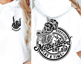 Könnte beinhalten: Weißer Kapuzenpullover mit schwarzen Grafikdesigns. Auf der Vorderseite befindet sich eine Skeletthand in einer Rock'n'Roll-Geste mit dem Wort "Motherhood". Auf der Rückseite ist ein Skelett mit Dutt zu sehen, das ein Weinglas hält, mit dem Text "Surviving Motherhood One Sip At A Time".