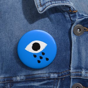 May include: A circular pinback button featuring a bright blue background with a central white eye graphic that has a solid black pupil and three black teardrops falling from the bottom edge of the eye, attached to a blue denim fabric surface.