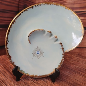 Può includere: Un posacenere in ceramica blu chiaro con un bordo dorato e un simbolo massonico al centro. Il simbolo è un compasso e una squadra con una "G" al centro.