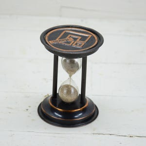 Op de afbeelding: Zwarte en bronzen zandloper met een cirkelvormige boven- en onderkant. De bovenkant heeft een bronzen embleem. De zandloper heeft een helder glazen midden met zand. Het frame is zwart met bronzen accenten. De zandloper is een decoratief item.