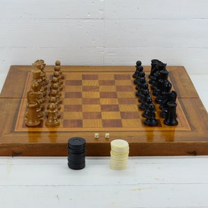 Puede incluir: Un tablero de ajedrez de madera con un juego de piezas de ajedrez negras y marrones. El tablero está abierto y las piezas están dispuestas para una partida. También hay dos pilas de damas negras y blancas al lado del tablero.