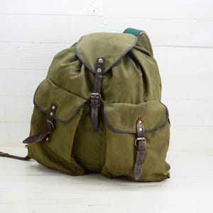 Op de afbeelding: Olijfgroene canvas rugzak met bruine leren riemen en gespen. De rugzak heeft twee zijzakken en een bovenklep. De tas is ontworpen voor gebruik buitenshuis en heeft een vintage uitstraling.
