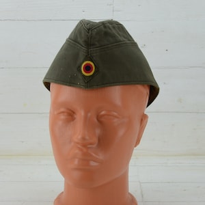 Könnte beinhalten: Eine grüne Militärkappe mit einem schwarzen, gelben und roten kreisförmigen Emblem auf der Vorderseite.