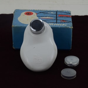 Può includere: Bottiglia di plastica bianca con un tappo argentato e un'etichetta bianca con un logo. La bottiglia è appoggiata su una superficie marrone scuro con una scatola blu dietro. La scatola ha un'etichetta bianca con il testo "СОВЕТСКАЯ МЕХАНИЧЕСКАЯ СВЕЧА" e un cerchio rosso con la lettera "E" all'interno.