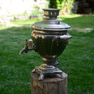 May include: A silver samovar with a spout and two handles, sitting on a wooden stump. The samovar is a traditional Russian tea kettle.