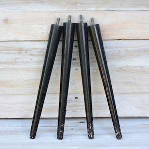 May include: Four black, tapered furniture legs with metal rods at the top. The legs are angled and appear to be made of wood. The legs are set against a light-colored, wooden background.