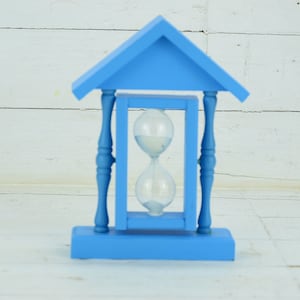 May include: A blue wooden hourglass with a white sand timer inside. The hourglass is in a blue wooden frame with a roof.