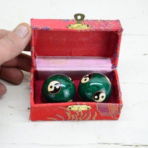 Può includere: Un paio di sfere cinesi verdi per la salute con simboli yin e yang, in una scatola rossa fantasia. La scatola ha una chiusura dorata e un interno rosa, suggerendo un focus sul benessere e sulle pratiche tradizionali.