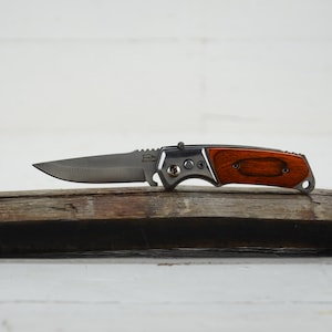 May include: A silver pocket knife with a wooden handle. The handle is a dark brown wood with a lighter brown wood inlay. The knife is closed and sitting on a piece of weathered wood.