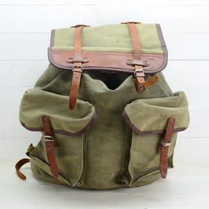 Puede incluir: Mochila de estilo vintage en lona verde oliva con detalles en cuero marrón. Cuenta con dos bolsillos laterales, correas de cuero y cierre de solapa. Diseñada para transportar objetos.