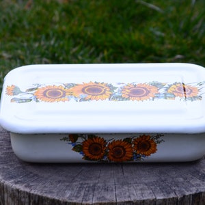 May include: White enamel baking dish with a sunflower design. The dish has a lid and is sitting on a wooden surface.