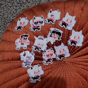 May include: A collection of 12 white vinyl stickers featuring cartoon characters with red, black, and white details. The characters are all different and have various expressions. The stickers are on a brown surface.