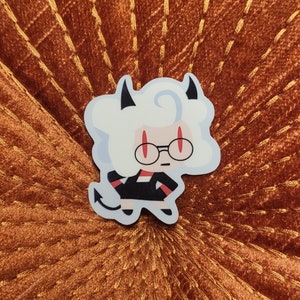 May include: A sticker featuring a cartoon character with white fluffy hair, black horns, and red-tinted glasses. The character is wearing a black outfit with red accents and a small tail. The sticker is placed on a textured orange surface.