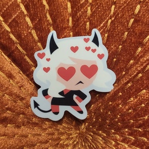May include: A white sticker with a cartoon character with red hearts for eyes. The character is wearing a black dress and has white hair. The sticker is on a brown background.