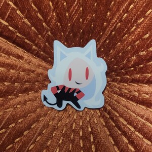 May include: A white and red cartoon character with a devil tail and horns. The character is wearing a red and black striped shirt and has red eyes. The character is on a brown background.
