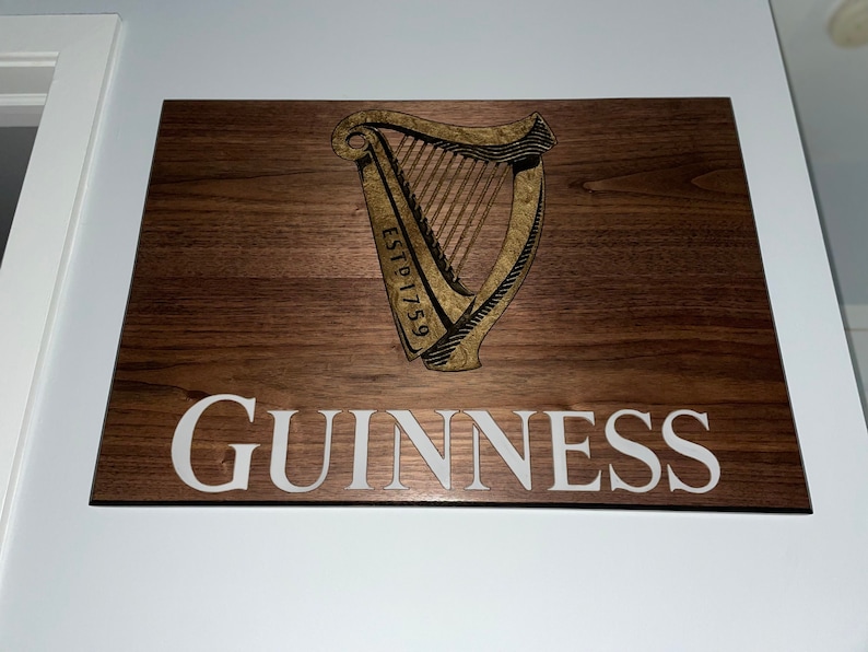 Pu&ograve; includere: Un cartello in legno con il logo Guinness, che presenta un'arpa dorata con il testo "ESTD 1759" e la parola "GUINNESS" in lettere bianche.