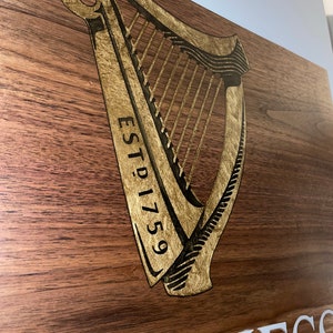 Pu&ograve; includere: Un cartello di legno con un'arpa dorata e il testo "ESTD 1759" e la parola "GUINNESS" in lettere bianche.