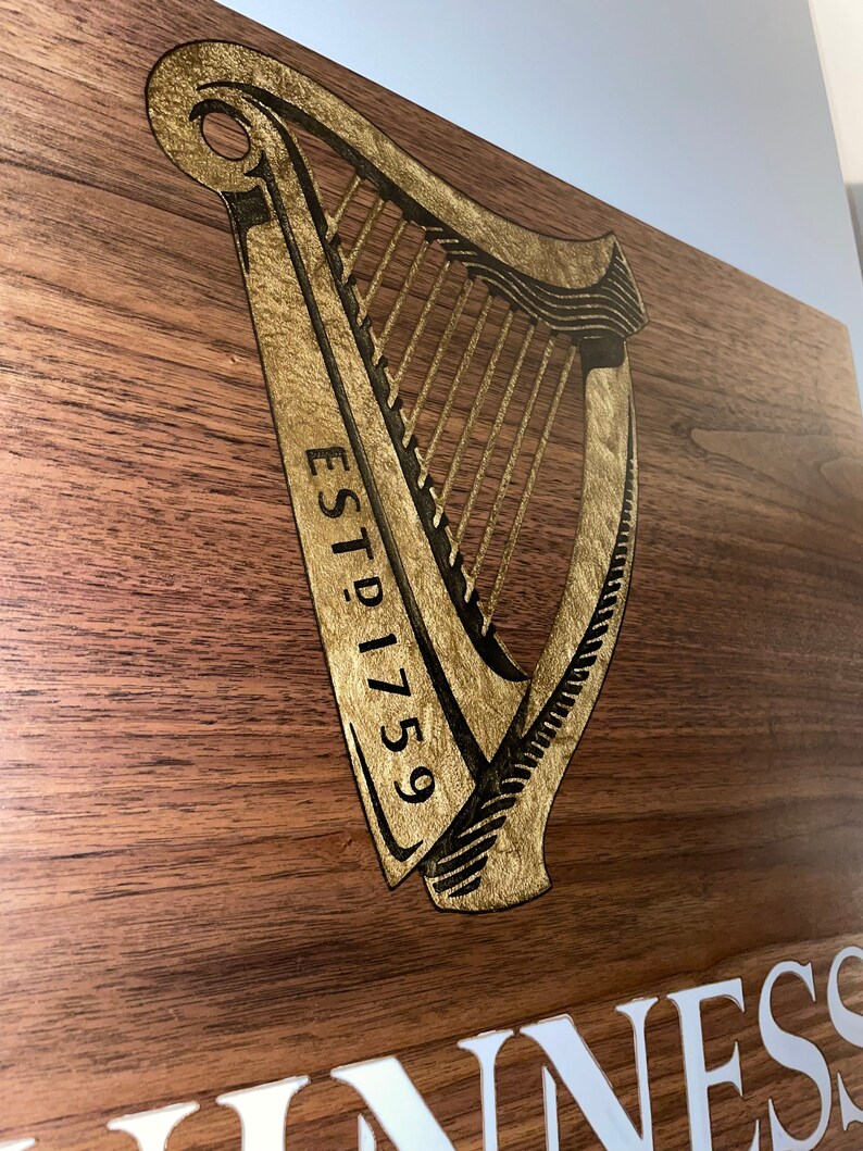 Pu&ograve; includere: Un cartello di legno con un'arpa dorata e il testo "ESTD 1759" e la parola "GUINNESS" in lettere bianche.