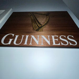 Pu&ograve; includere: Un cartello in legno con la parola "GUINNESS" in lettere bianche e un logo di arpa dorato con il testo "ESTD 1759".
