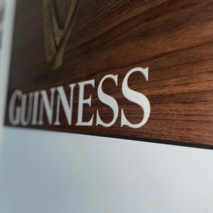 Pu&ograve; includere: Un cartello di legno con la parola "GUINNESS" in lettere bianche. Il cartello ha una venatura del legno marrone scuro.