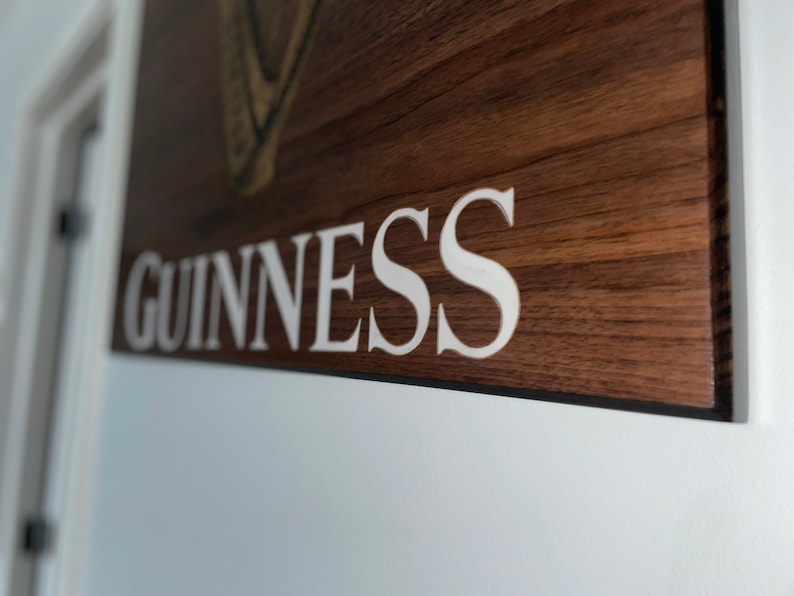 Pu&ograve; includere: Un cartello di legno con la parola "GUINNESS" in lettere bianche. Il cartello ha una venatura del legno marrone scuro.