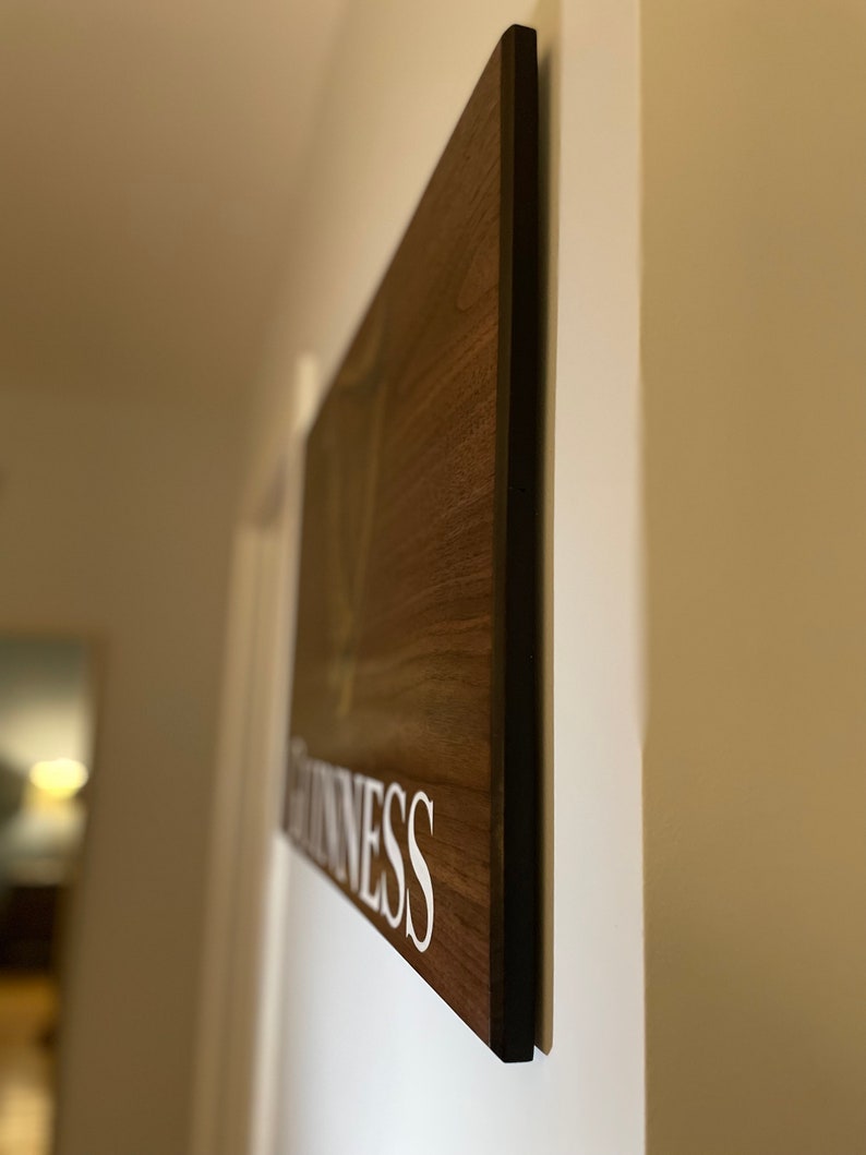 Pu&ograve; includere: Un cartello di legno con la parola "GUINNESS" stampata in lettere bianche su uno sfondo marrone. Il cartello &egrave; montato su una parete bianca.