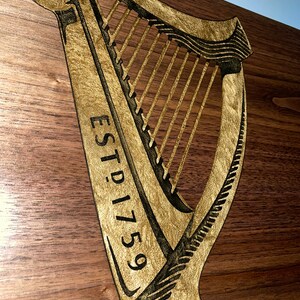 Pu&ograve; includere: Un cartello di legno con un'arpa dorata e il testo "ESTD 1759" inciso su di esso.